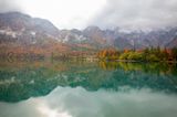 Weiter Blick über den See auf eine kleine Hütte in der Bildmitte, umgeben von herbstlichen Wäldern und felsigen Bergspitzen