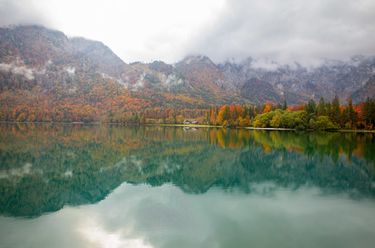 Weiter Blick über den See auf eine kleine Hütte in der Bildmitte, umgeben von herbstlichen Wäldern und felsigen Bergspitzen