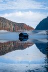 Blick über den See auf eine Fischerhütte, links daneben ein Waldhang herbstlich angeleuchtet. Rechts ein Berghang und Wald im Schatten.